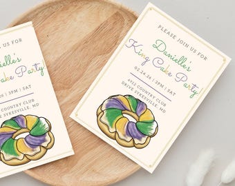 May include: Two cream-coloured invitations with gold borders and a watercolour illustration of a King Cake. The text reads "Please join us for Danielle's King Cake Party!" with date, time, and location details. The invitations are on a wooden tray.