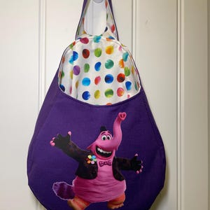 May include: A purple tote bag featuring a cartoon character with a pink body, black jacket, and a bow tie. The bag's interior lining has a white background with colorful polka dots. The bag is hanging on a white hook.