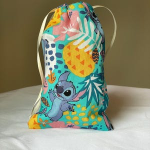 May include: A drawstring bag featuring a colorful tropical pattern with a cartoon character. The bag's design includes a teal background with pink, yellow, and white accents, and a light blue character. The bag has a cream-colored drawstring.