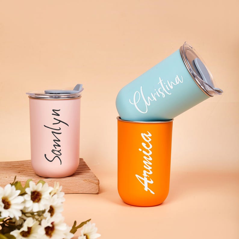 May include: Three cylindrical tumblers with lids, one light pink with the name Samlyn, one light blue with the name Christina, and one orange with the name Arnica. Each tumbler features a smooth matte finish and white script lettering.