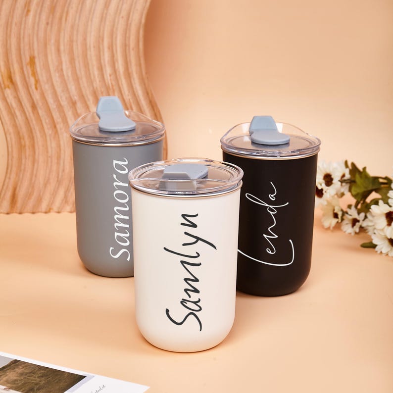 May include: Three cylindrical tumblers with clear lids and light blue stoppers. The gray tumbler features the name Samora, the white tumbler features the name Samlyn, and the black tumbler features the name Lenda in white cursive script lettering.