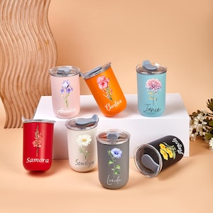 May include: A collection of insulated travel mugs in various colors, each adorned with a floral design and a personalized name. The mugs feature a lid and are displayed on a white surface against a neutral backdrop. The colors include pink, orange, blue, red, white, gray, and black.