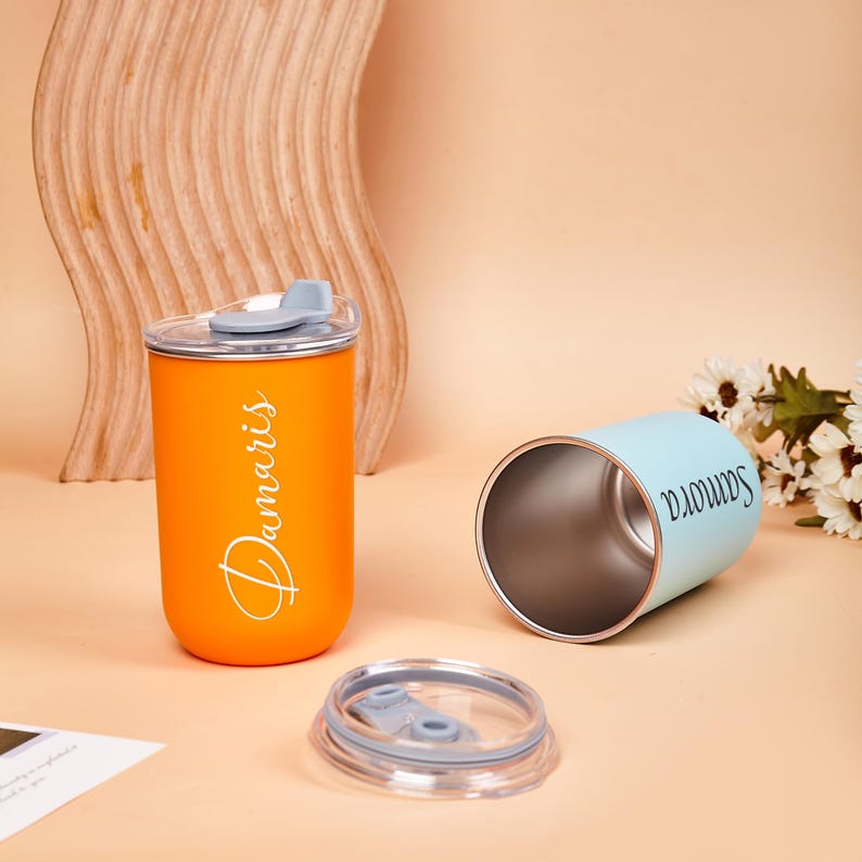 May include: An orange tumbler with a gray lid and the name Damaris printed in white, alongside a light blue tumbler with the name Samora printed in black, and a separate clear plastic lid with a gray sliding closure mechanism.