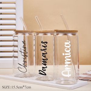 May include: Three clear glass drinking cups with bamboo lids and glass straws. Each cup has a name printed on it: Christina, Damaris, and Arnica. The cups are on a white tray. Size: 15.5cm x 7cm.