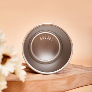 May include: A circular stainless steel cup viewed from above, featuring a smooth metallic interior finish. The base of the cup is clearly embossed with the text SUS 304, indicating the material grade of the metal construction.