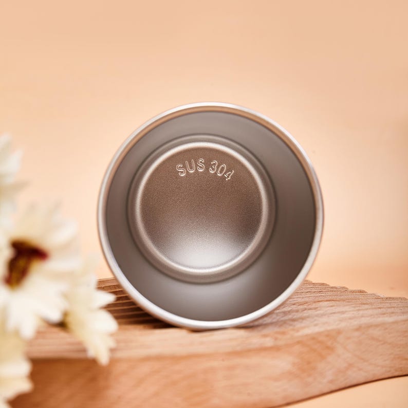 May include: A circular stainless steel cup viewed from above, featuring a smooth metallic interior finish. The base of the cup is clearly embossed with the text SUS 304, indicating the material grade of the metal construction.