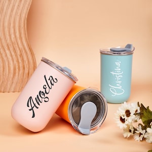 May include: Three cylindrical tumblers with clear lids and gray sliding closures. One is light pink with the name Angela in black script, one is light blue with the name Christina in white script, and one is solid orange with a metallic interior.