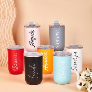 May include: Seven cylindrical tumblers with clear lids, each featuring a unique name in script lettering. The tumblers come in various matte colors including red, black, orange, light blue, white, grey, and light pink, arranged on a flat surface.