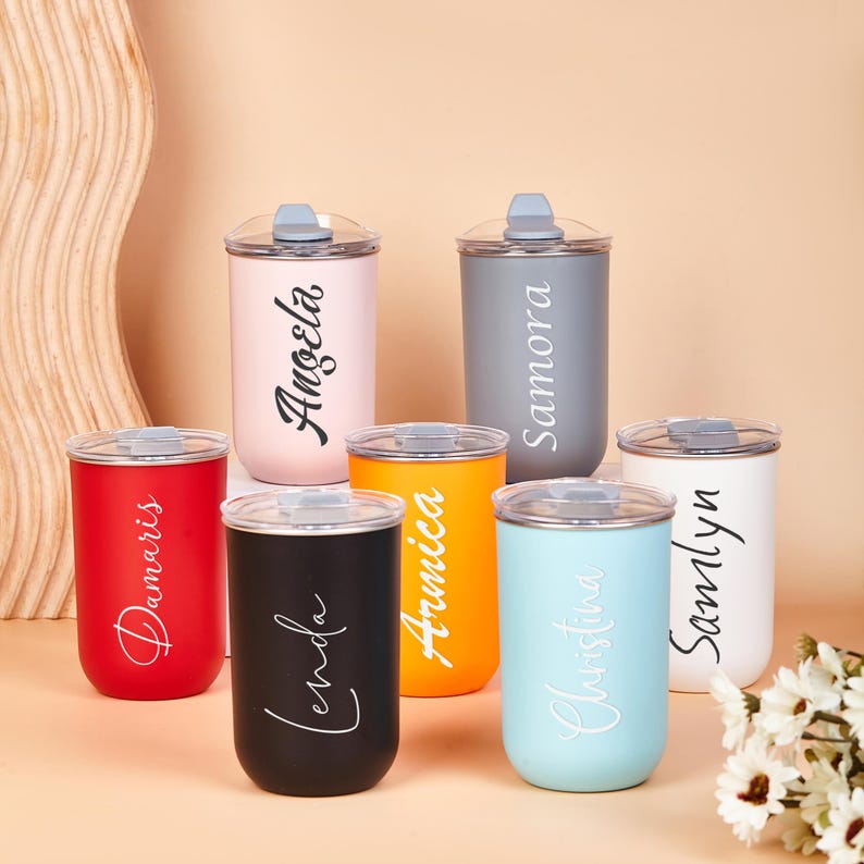 May include: Seven cylindrical tumblers with clear lids, each featuring a unique name in script lettering. The tumblers come in various matte colors including red, black, orange, light blue, white, grey, and light pink, arranged on a flat surface.