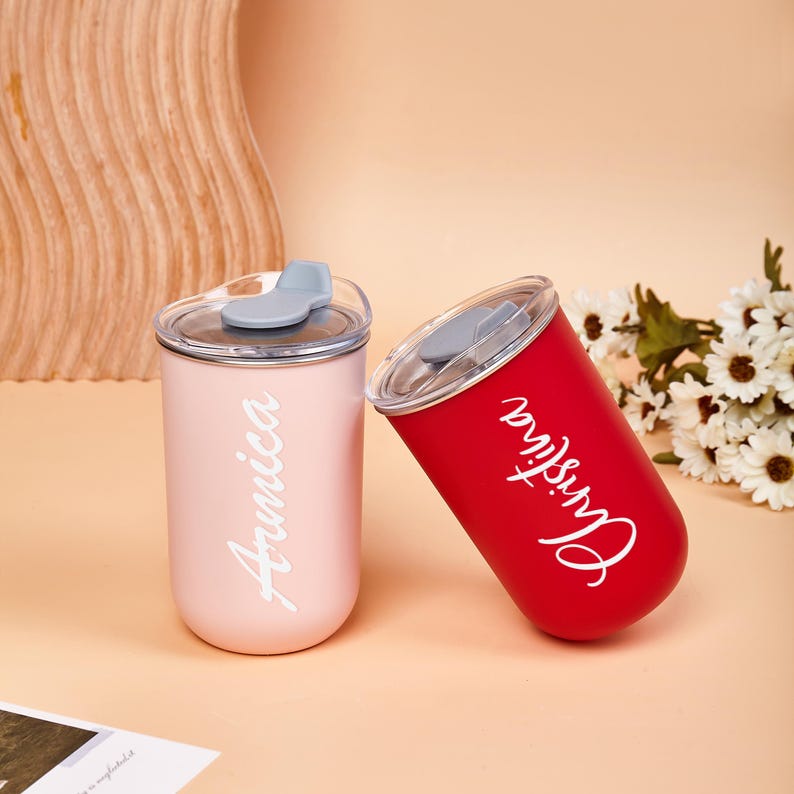 May include: Two cylindrical tumblers with clear lids and grey sliding closures. One tumbler is matte light pink with the white cursive name Arnica, and the other is matte red with the white cursive name Christina printed vertically on the side.