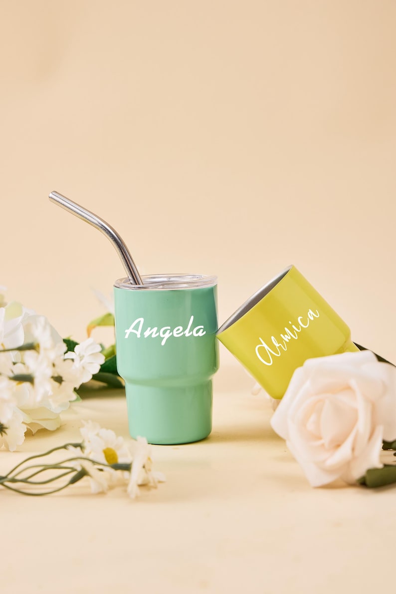May include: Two personalized tumblers, one mint green and one yellow, with white text. The mint green tumbler has a silver straw. The text on the mint green tumbler says "Angela", and the yellow tumbler says "Armica".