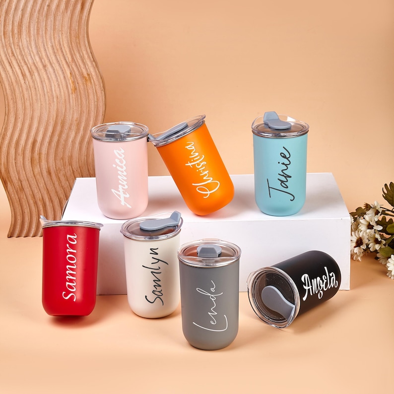 May include: Seven cylindrical tumblers in various colors including red, light pink, orange, light blue, white, gray, and black. Each tumbler features a clear lid and a personalized name printed in a script font: Samora, Annica, Christina, Janie, Samlyn, Lenda, and Angela.