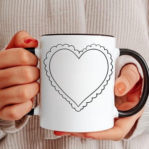 May include: White ceramic mug with a black handle and rim, featuring a large heart design outlined in black. The heart is surrounded by a scalloped border. The mug is held by a person.