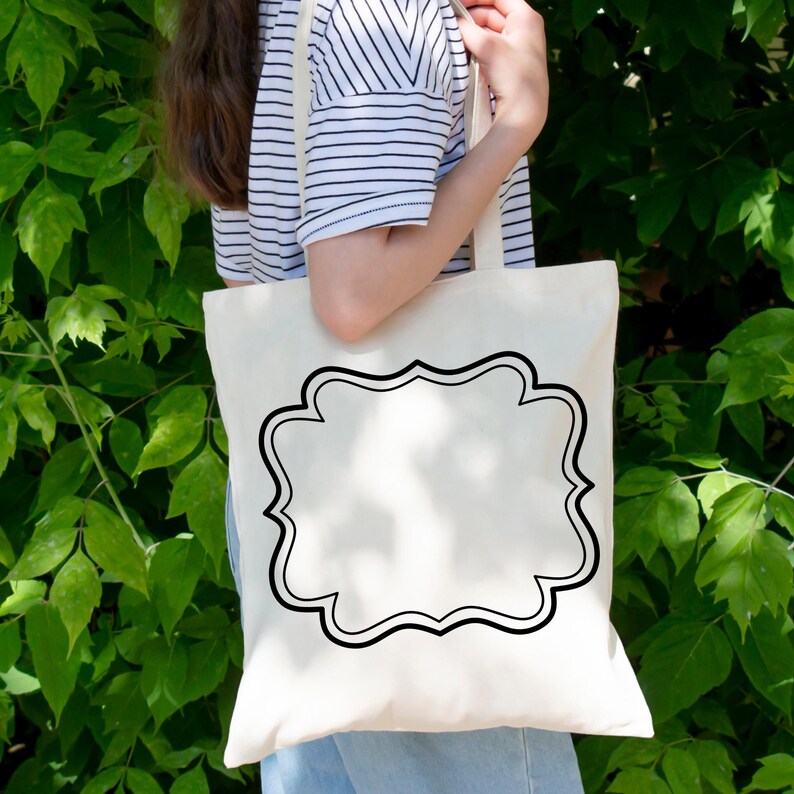 May include: Cream-colored tote bag with a black decorative frame design. The bag is made of a sturdy fabric and has a simple, elegant design. The bag is held by a person in a striped shirt.