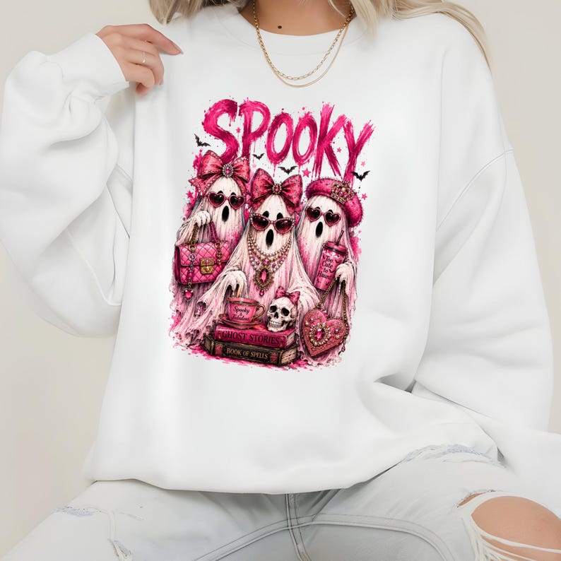 May include: A white long-sleeve sweatshirt featuring a graphic print of three ghosts wearing pink bows and heart-shaped sunglasses. The text SPOOKY appears above the ghosts, who are surrounded by pink accessories, books, and a skull.