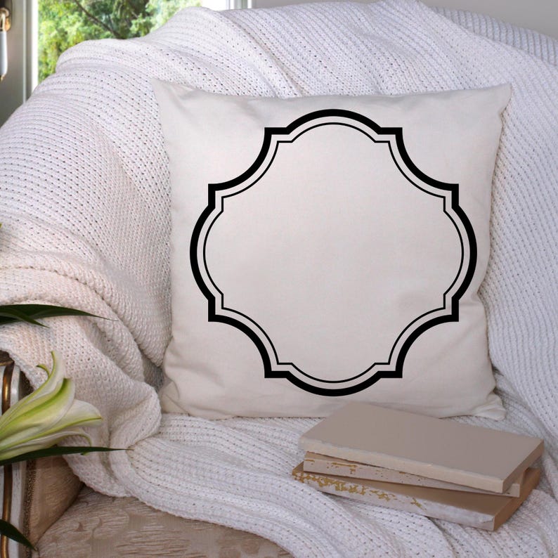 May include: A white decorative pillow with a black quatrefoil design. The pillow is on a white knitted throw, with a stack of books and a lily flower in the background. The pillow is square-shaped.