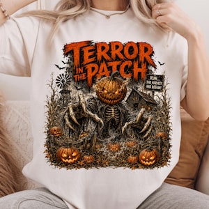 May include: A cream-colored t-shirt featuring a graphic print of a skeletal creature with a pumpkin head in a field. The text reads "TERROR IN THE PATCH" and "RUN... THE HARVEST HAS BEGUN". The design includes a windmill, a barn, and several jack-o'-lanterns.