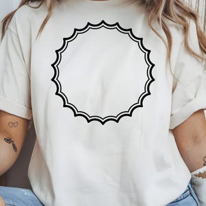 May include: Cream-colored t-shirt featuring a large, black, scalloped circle design. The design is centered on the shirt, creating a simple yet eye-catching graphic. The shirt has short sleeves and a relaxed fit, suitable for casual wear.