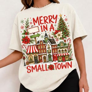 May include: A cream-colored t-shirt featuring a festive graphic print of a small town during the holidays. The design includes red text that reads MERRY IN A SMALL TOWN, a decorated Christmas tree, poinsettias, candy canes, and buildings.