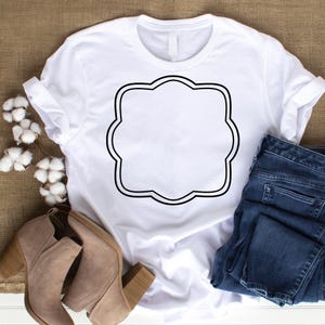 May include: A white t-shirt with a black scalloped frame design. The shirt is paired with blue jeans and tan ankle boots. The design is centered on the shirt, offering a blank space for customization or a graphic.