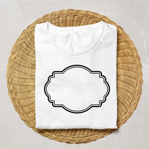 May include: A folded white t-shirt with a black decorative label design. The shirt is placed on a round, woven wicker placemat. The label is a double-lined, ornate shape, ready for custom text or design.