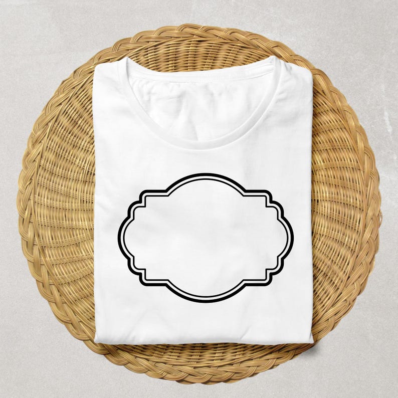 May include: A folded white t-shirt with a black decorative label design. The shirt is placed on a round, woven wicker placemat. The label is a double-lined, ornate shape, ready for custom text or design.
