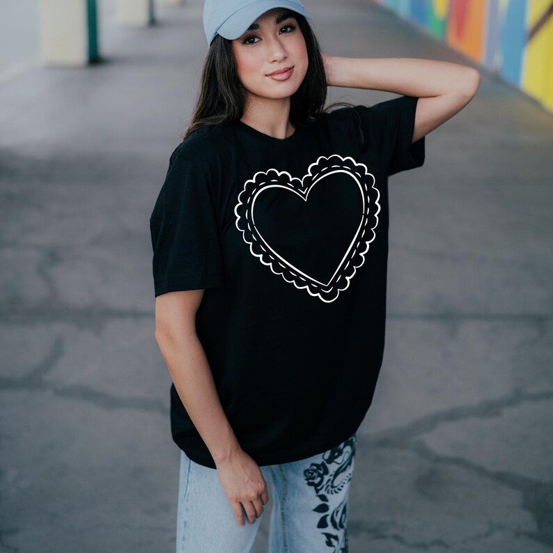 May include: Black t-shirt featuring a white heart design with a scalloped border. The heart is centered on the shirt. The shirt is made of a soft material. The model is wearing a light blue baseball cap and light blue jeans.