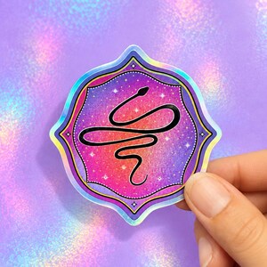 May include: A die-cut sticker featuring a black line-art snake coiled in a horizontal S-shape. The background is a vibrant gradient of pink, purple, and orange with a holographic finish, decorated with small white stars inside a scalloped frame.