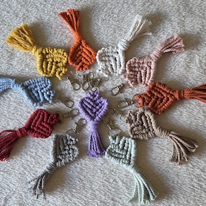 Peut inclure: Une collection de onze porte-clés en macramé en forme de cœur disposés en cercle. Chaque porte-clé est fabriqué à partir de cordon en coton dans diverses couleurs, notamment jaune, orange, blanc, rose, bleu, rouge, violet, gris, vert clair, beige et rouille.