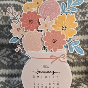 May include: A floral-themed 2026 calendar featuring a vase shape with a bouquet of paper flowers in shades of pink, yellow, and peach. The calendar displays the month of January with the days of the week and dates. The calendar is approximately 13 cm tall.
