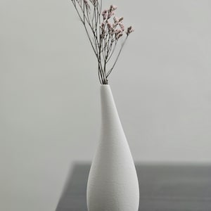 Könnte beinhalten: Eine weiße, strukturierte Vase in Tropfenform, gefüllt mit zarten Trockenblumen mit kleinen, rosa Blüten. Die Vase steht auf einer dunklen Oberfläche, was einen Kontrast erzeugt. Das Design ist minimalistisch und elegant, passend für die Inneneinrichtung.