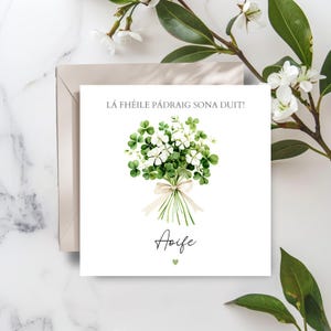 May include: A white greeting card featuring a watercolour illustration of a bouquet of shamrocks and white flowers, tied with a cream ribbon. The text "LÁ FÉILE PÁDRAIG SONA DUIT!" is at the top, with the name "Aoife" below the bouquet.