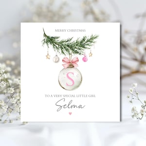 May include: A festive Christmas card with a watercolour design. The card features a bauble with the letter "S", a pink bow, and the text "Merry Christmas" and "To a very special little girl Selma".