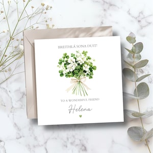 May include: A white greeting card featuring a watercolour illustration of a bouquet of white flowers and green shamrocks. Text reads "Breithlá Sona Duit!" and "To a Wonderful Friend Helena". The card is paired with a beige envelope.
