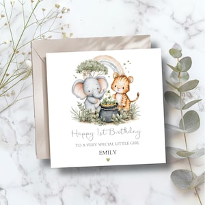 May include: A square birthday card featuring an elephant and tiger cub illustration. The card reads "Happy 1st Birthday" with the text "To a very special little girl" and the name "EMILY". A rainbow and tree are in the background, with a beige envelope.