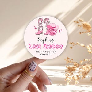 Könnte beinhalten: Ein runder Aufkleber mit rosa Hintergrund, der eine Illustration von Cowboy-Stiefeln und -Hut zeigt. Der Text lautet "Sophia's Last Rodeo" und "Thank you for coming!" in Rosa. Der Aufkleber wird von einer Hand mit einem goldenen Ring gehalten.