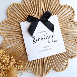 May include: A white greeting card with the text "TO A SPECIAL Brother In-Law" and the name "Edgar" with "ON YOUR BIRTHDAY" underneath. The card is adorned with a black satin bow. The card rests on a woven placemat.