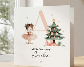 Puede incluir: Una tarjeta de felicitación cuadrada con una letra A grande de color rosa pálido, una bailarina con tutú rosa y un árbol de Navidad decorado con regalos. El texto dice Merry Christmas Amelia en fuente negra con un pequeño corazón rosa.
