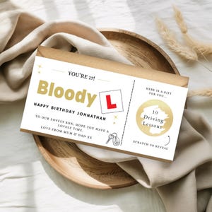 May include: A birthday card with the text "Bloody L" and "Happy Birthday Johnathan". The card includes a scratch-off area for 10 driving lessons and the message "Love from Mum & Dad XX". The card is on a wooden plate.