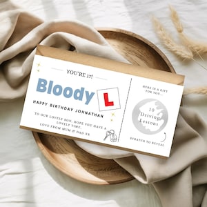 May include: A birthday gift card with the text "Bloody L" and "Happy Birthday Johnathan". The card features a red "L" symbol, a key graphic, and a scratch-off area for 10 driving lessons. The card is presented on a wooden plate.