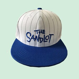 Può includere: Cappellino da baseball bianco e blu con la scritta "THE SANDLOT" ricamata in blu. Il cappellino ha una visiera blu e una corona bianca con sottili gessati neri verticali. Un bottone blu è in cima.