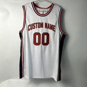 Puede incluir: Camiseta de baloncesto blanca con detalles rojos y azul marino. La camiseta presenta el texto "CUSTOM NAME" sobre el número "00" en rojo. La camiseta tiene un diseño de malla y está colgada de una percha metálica.