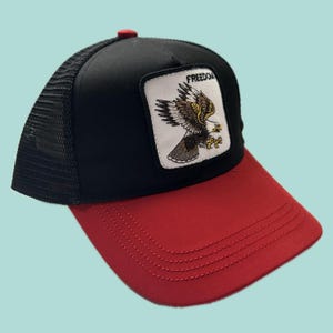 Può includere: Cappellino da camionista nero e rosso con retro in rete e visiera curva. Il pannello frontale presenta una toppa con un'aquila e la scritta "FREEDOM". Il cappello ha un bottone rosso sulla parte superiore.