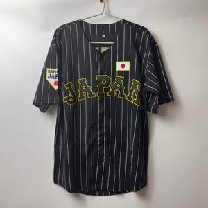Op de afbeelding: Zwart honkbalshirt met witte verticale strepen. Het woord "JAPAN" staat in gele letters over de borst. Een Japanse vlagpatch staat op de borst en een "SAMURAI JAPAN" patch op de linkermouw.