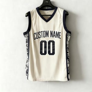 Op de afbeelding: Crèmekleurig basketbalshirt met een marineblauwe V-hals en bies. Het shirt toont de tekst "CUSTOM NAME" boven het nummer "00" in marineblauw. De zijkanten hebben een zwart-wit patroon.