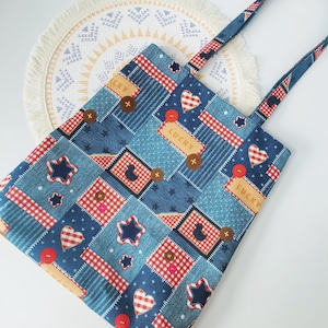 以下が含まれることがあります： 星、ハート、ボタンの繰り返し模様が特徴の青いパッチワークトートバッグ。バッグはデニムのような質感で、お揃いのストラップが付いています。「LUCKY」の文字が長方形のパッチにプリントされています。