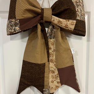 May include: A large decorative fabric bow featuring a patchwork design in shades of brown, tan, and cream. The bow incorporates various patterns including white polka dots, dark brown leaf prints, and intricate floral paisley motifs throughout.