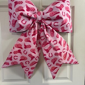 May include: A large, decorative bow in shades of pink, adorned with a repeating pattern of cowboy hats, boots, and horseshoes. The bow is hanging on a white door, adding a touch of country charm to any space.
