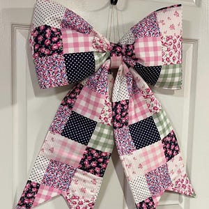 Puede incluir: Un gran lazo decorativo hecho de tela de retazos. El lazo presenta una variedad de patrones, incluyendo cuadros rosas, estampados florales, lunares y azul marino con puntos blancos. Cuelga de una puerta blanca, agregando un toque de encanto a la decoración.