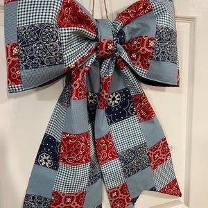 May include: A large decorative bow made of fabric featuring a patchwork design. The pattern consists of alternating squares in red, navy blue, and light blue, showcasing bandana-style paisley prints and small gingham check motifs throughout the piece.